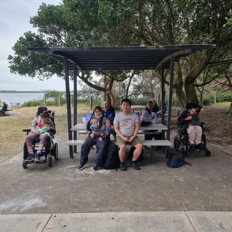 Day program participants and support workers relaxing at a picnic shelter by the water